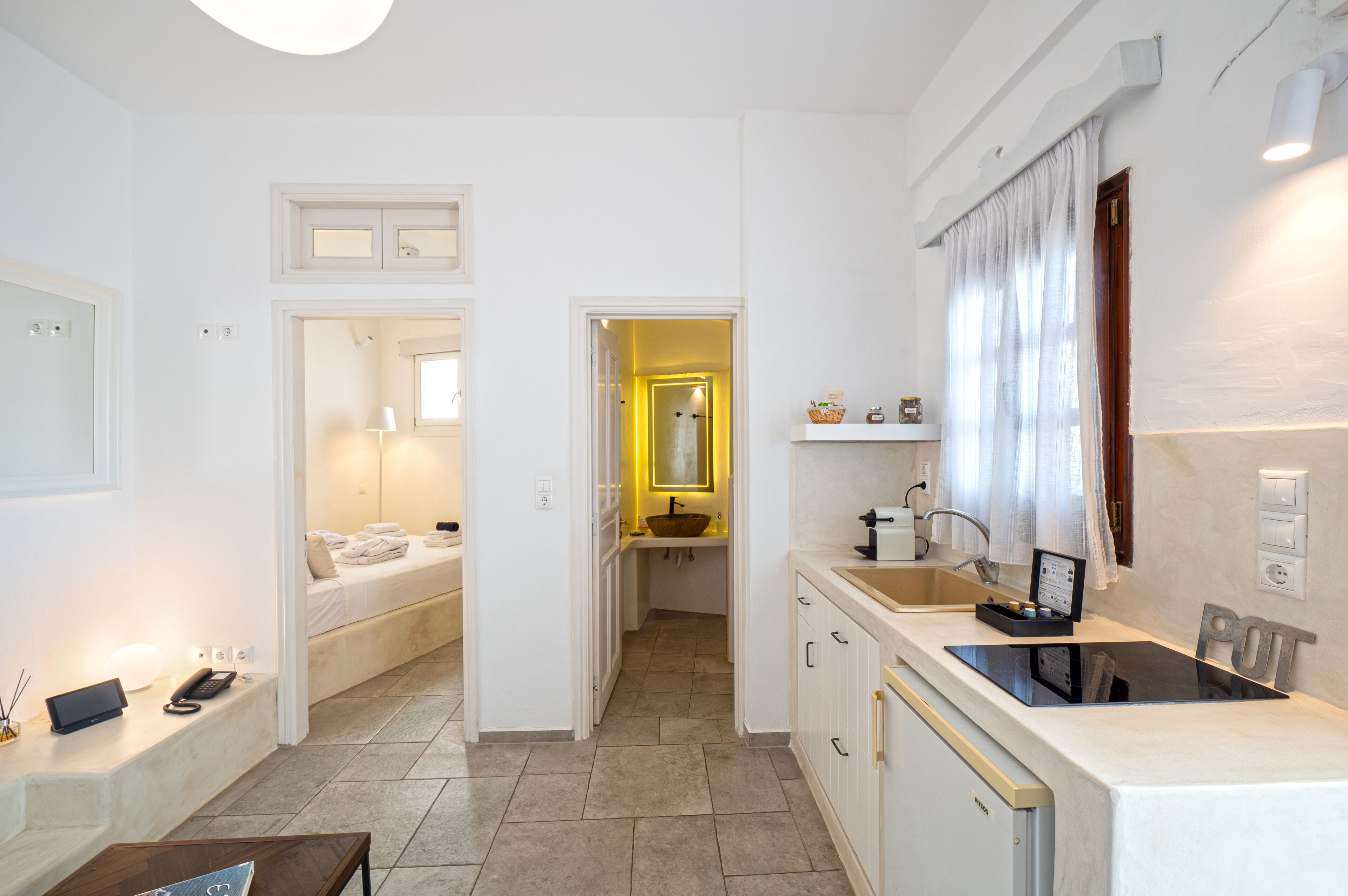 A compact hotel room with a bed, bedside table, and a small sitting area. It includes a kitchenette with a sink and a stove.