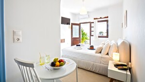 A hotel room with a bed, a bedside table with a lamp, a chair, a table with fruit and a newspaper, and a TV on the wall. - Kallichoron Art Hotel (Astypalaia)