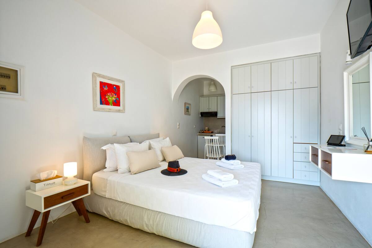 Une chambre d’hôtel moderne avec un lit soigneusement fait, une table de chevet avec une lampe, un petit bureau avec un ordinateur portable, et une vue sur la cuisine à travers une arche.