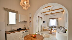 A modern kitchen with white cabinets, a wooden dining table with fruit, and a living area with a sofa and coffee table. - Kallichoron Art Hotel (Astypalaia)