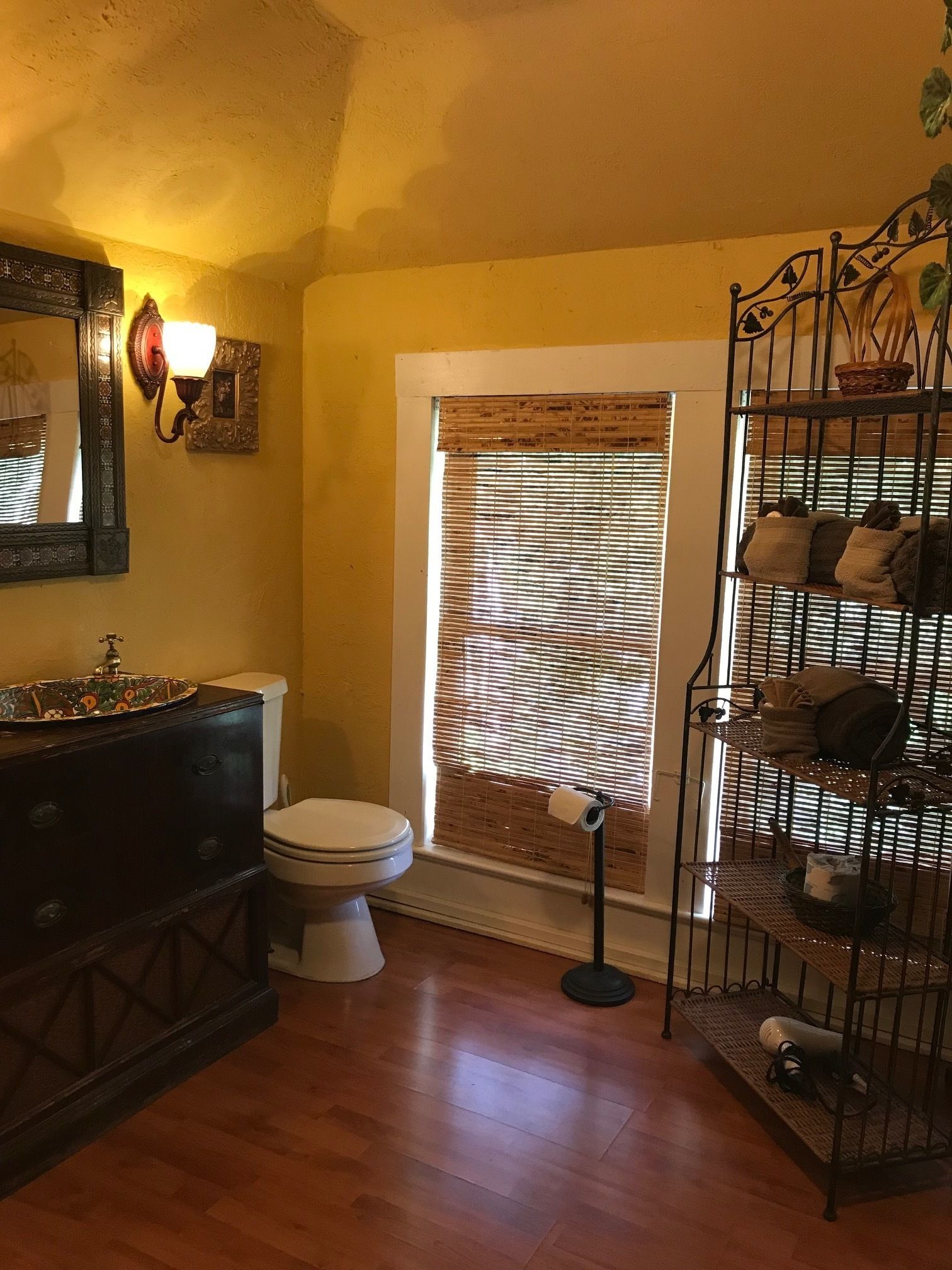 A bathroom with a toilet, a sink with a decorative basin, a mirror with a wall-mounted light, a window with blinds, and a metal rack with towels.