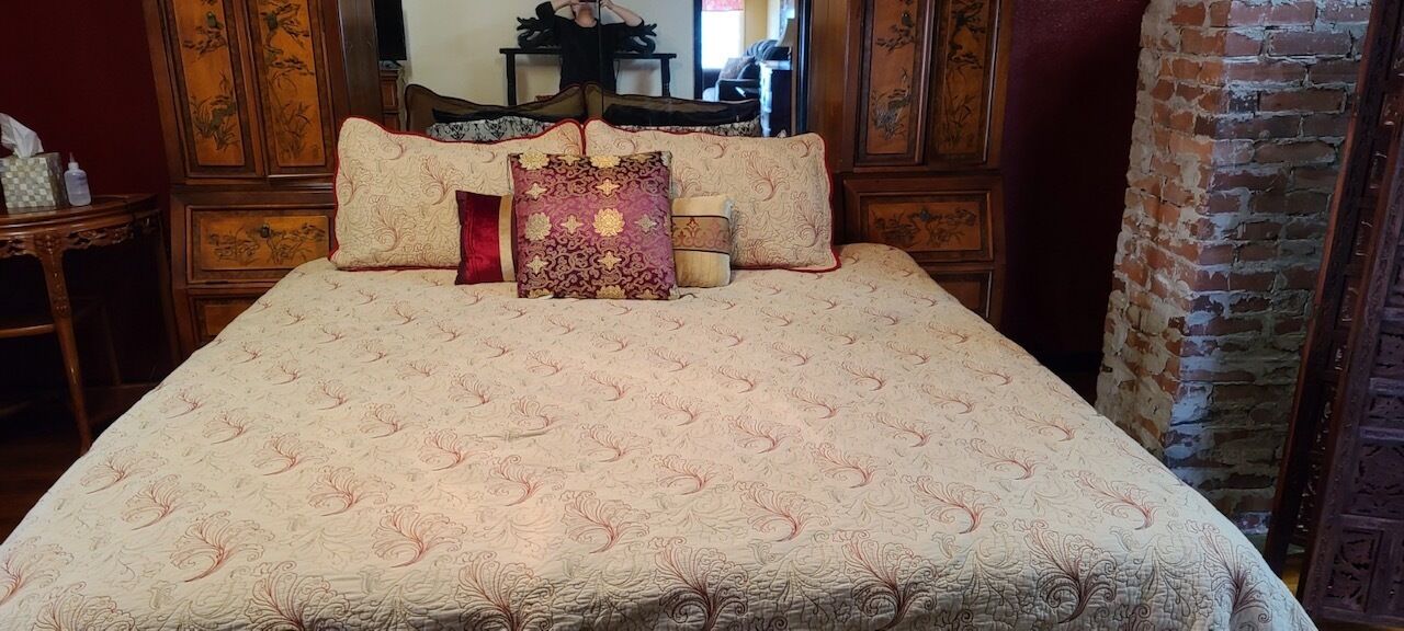 A bed with a patterned bedspread and multiple pillows, a wooden nightstand with a lamp, and a brick wall.