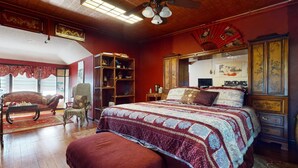 Ein traditionell eingerichtetes Zimmer mit einem großen Bett, einem Holzsessel, einem Bücherregal und einem Deckenventilator.