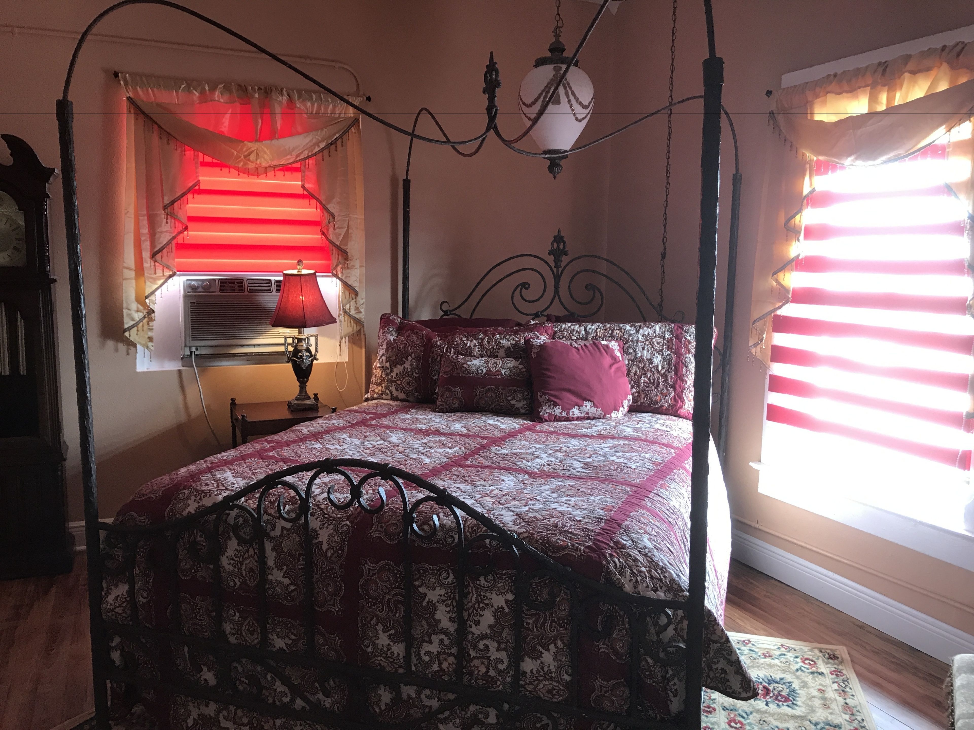 A bedroom with a four-poster bed, a bedside table with a lamp, a window with blinds, and a wall-mounted air conditioning unit.