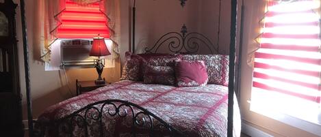 A bedroom with a four-poster bed, a bedside table with a lamp, a window with blinds, and a wall-mounted air conditioning unit.