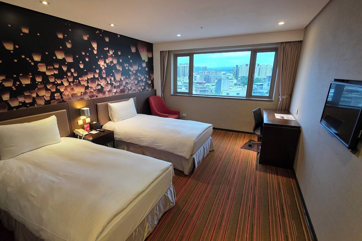 A hotel room with two beds, a desk, a chair, a TV, and a large window offering a city view.