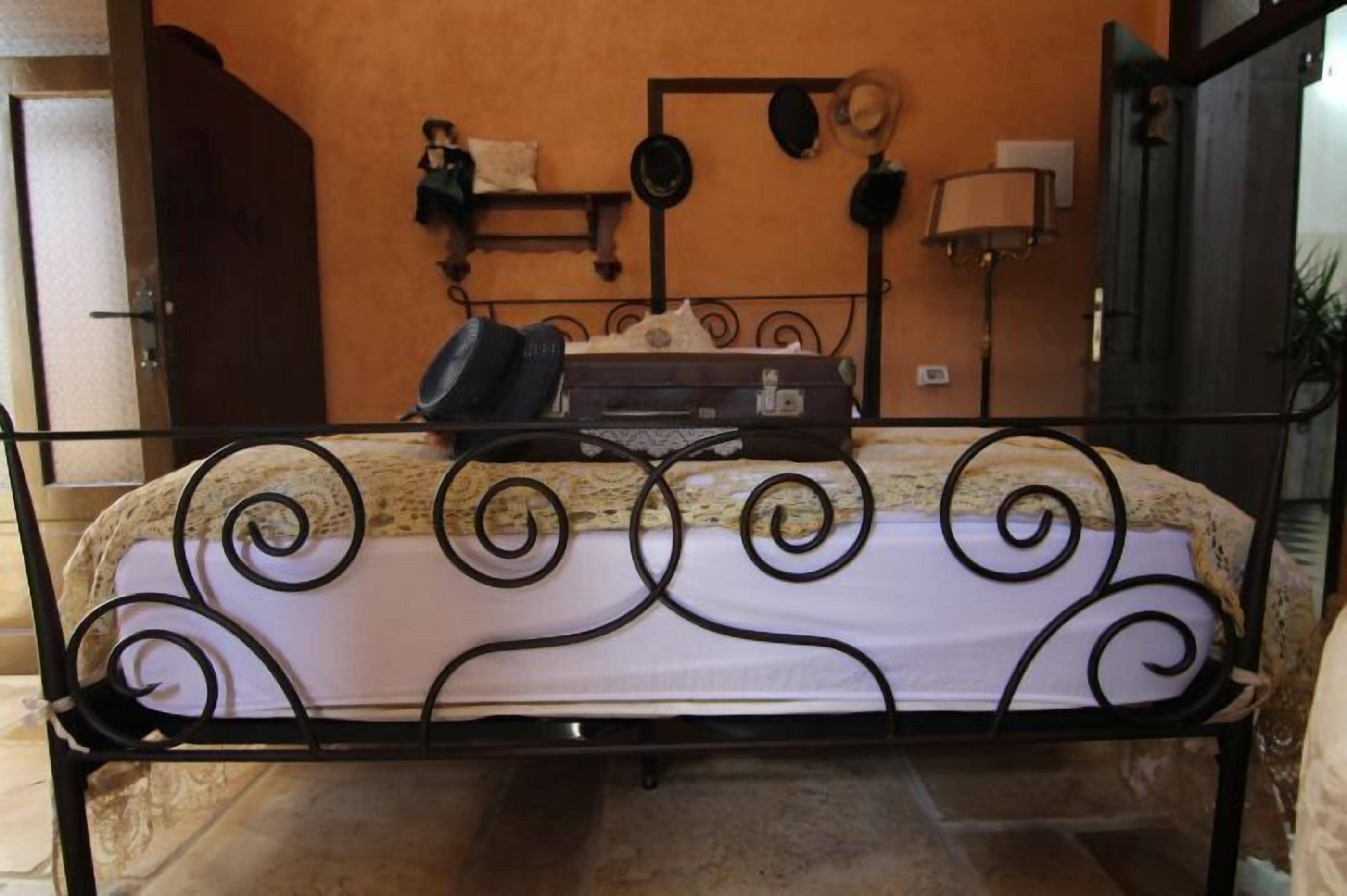 Una cama con estructura metálica y adornos en espiral, una maleta y un sombrero en el cabecero.