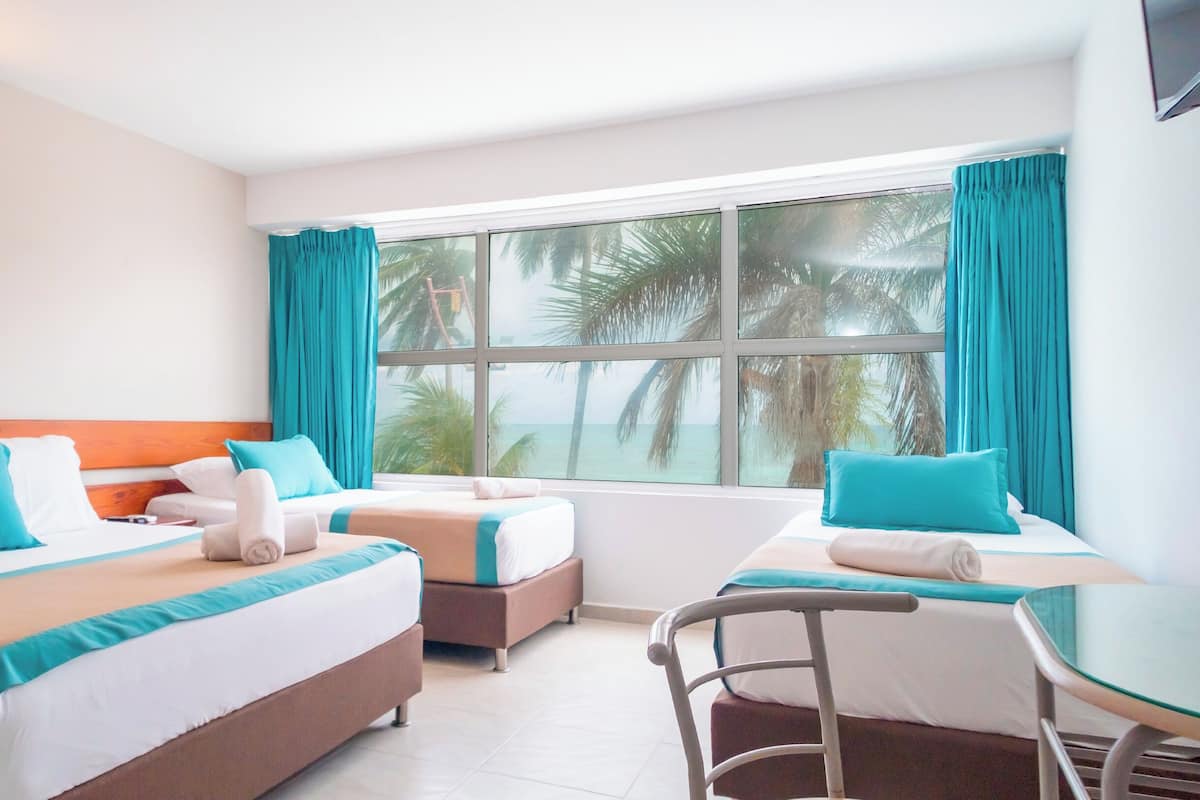 A hotel room with two beds, a glass table, and a view of palm trees and the ocean.