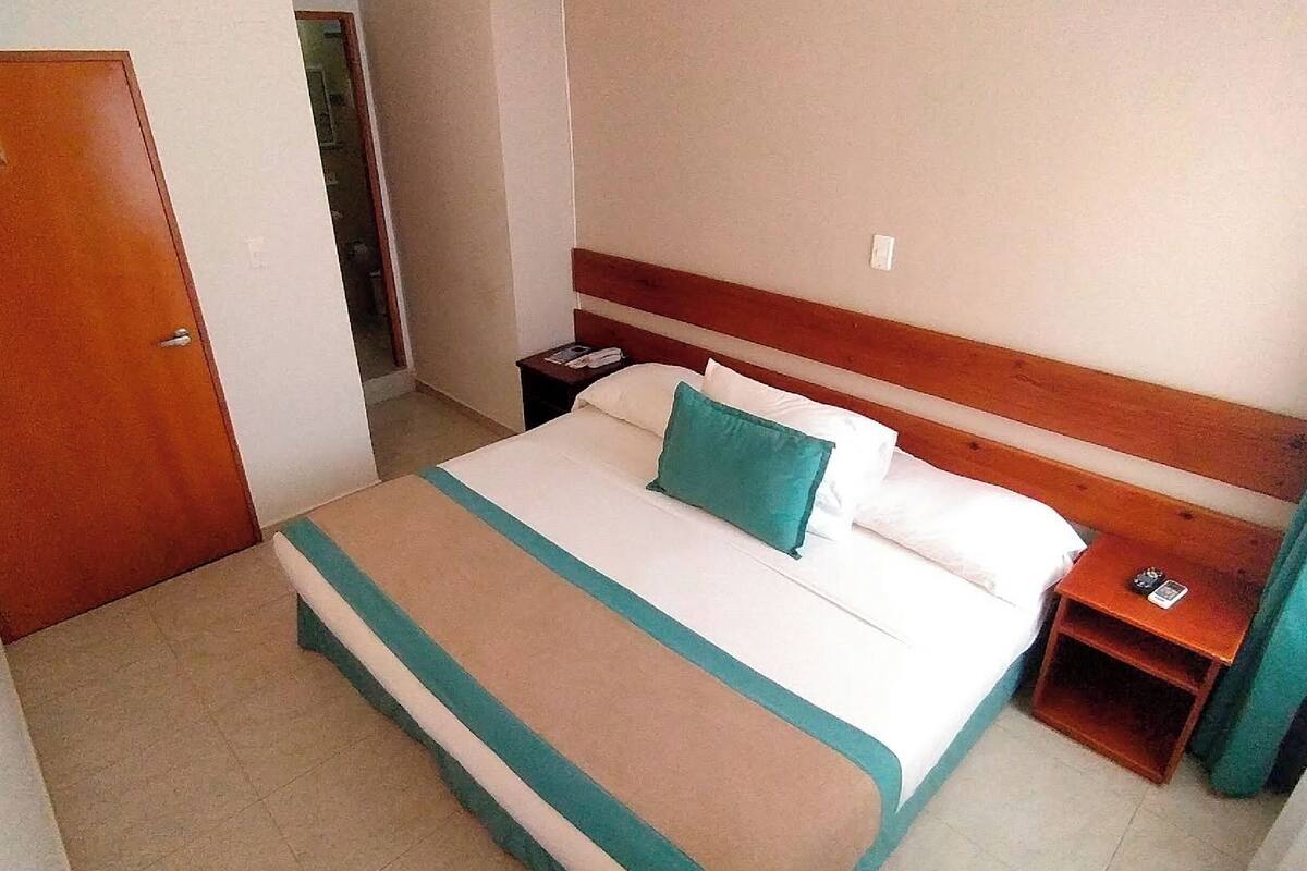 A hotel room with a bed, a nightstand, a wooden headboard, a door, and a curtain.