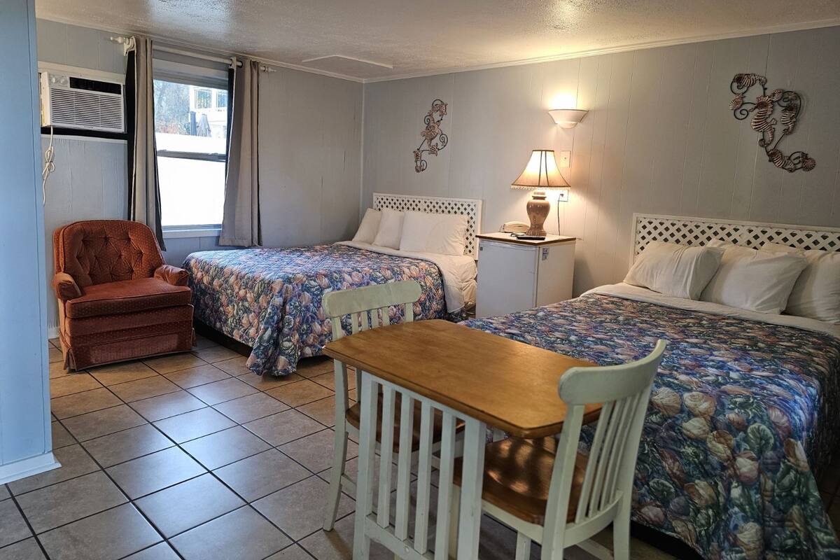 A room with two beds, a small table, a chair, a lamp, and a window with curtains.