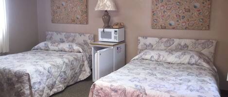 A room with two beds, each with floral-patterned bedding, a small refrigerator, a microwave, and a lamp.