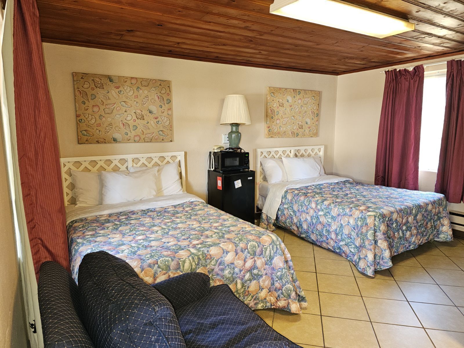 A room with two beds, each with floral bedding, a small refrigerator, a lamp, and artwork on the walls.