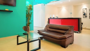 Eine moderne Lobby mit einem braunen Ledersofa, einem Glastisch, einem Blumentopf, einem roten Empfangsschalter und einem an der Wand befestigten Fernseher.