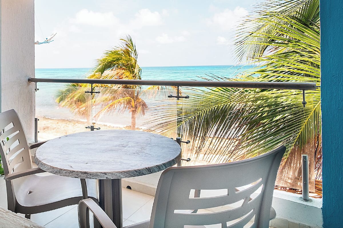 A balcony with a round table and two chairs, overlooking a beach with palm trees and the ocean.