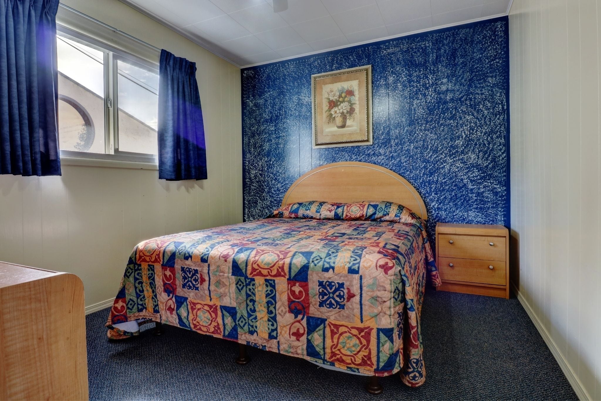 Une chambre à coucher avec un lit, une commode, une fenêtre avec des rideaux et un mur bleu texturé orné d’un tableau encadré.