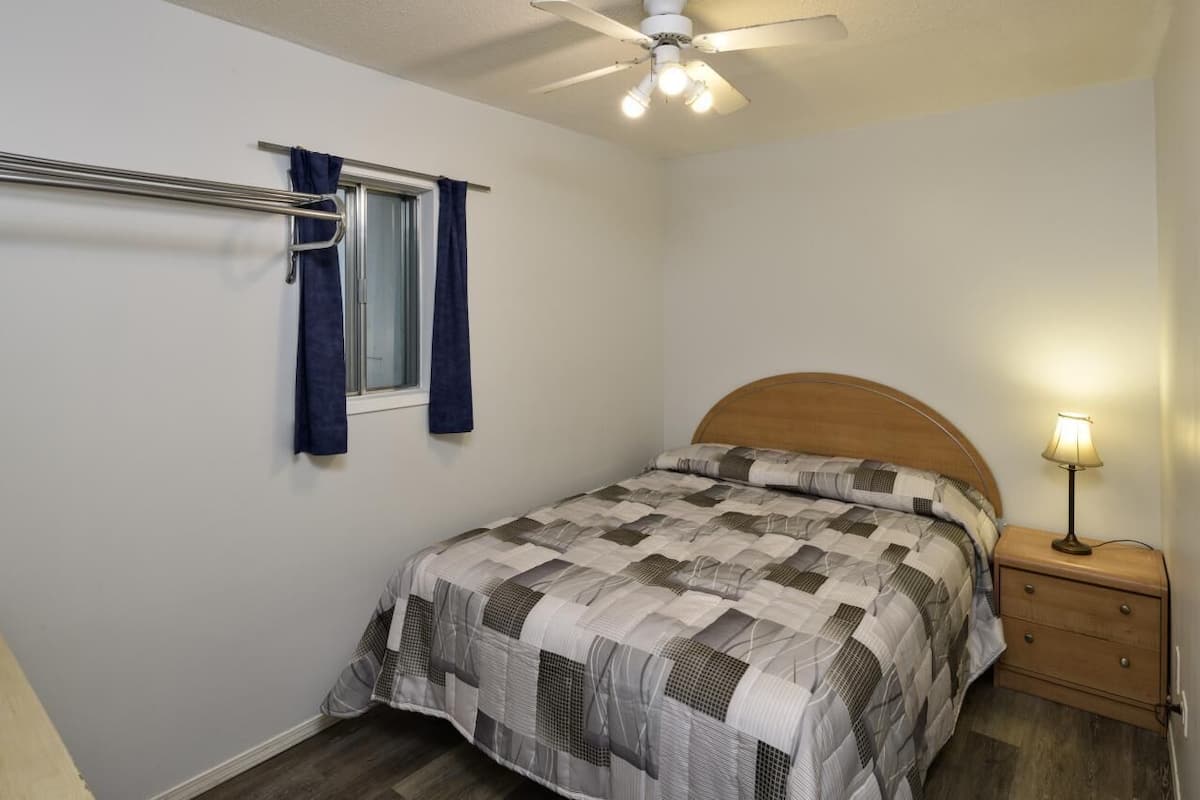 A bedroom with a bed, a nightstand, a lamp, a ceiling fan, and a window with curtains.