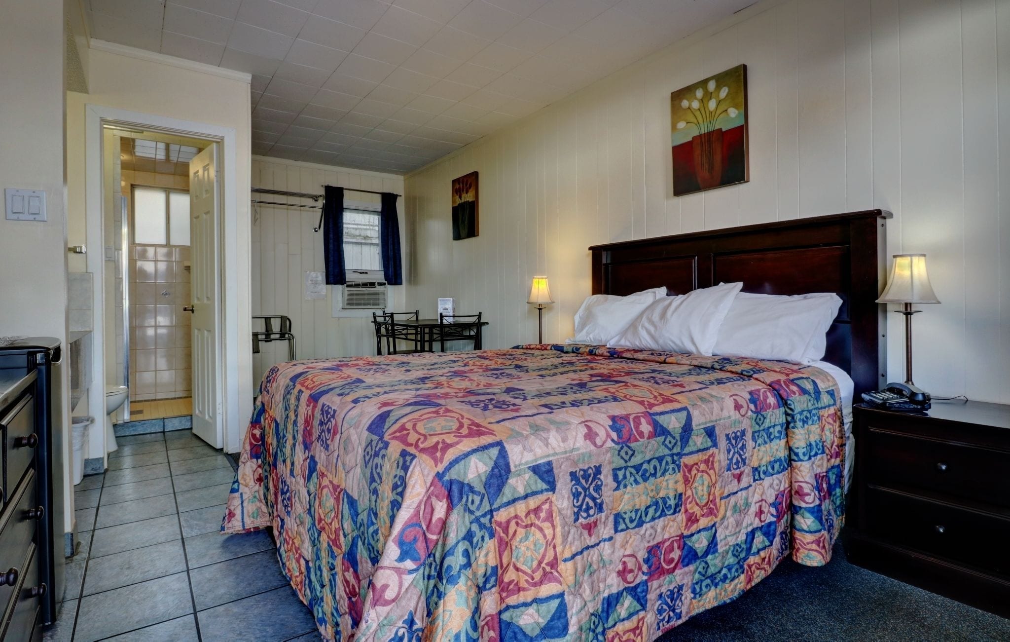 A hotel room with a bed, bedside table, lamp, and a desk with a chair. There is a bathroom with a shower and toilet visible through an open door.