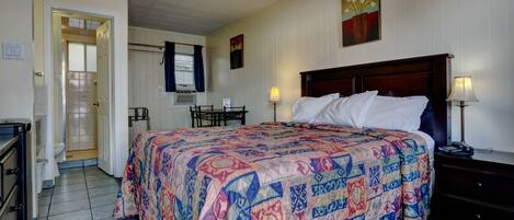 A hotel room with a bed, bedside table, lamp, and a desk with a chair. There is a bathroom with a shower and toilet visible through an open door.