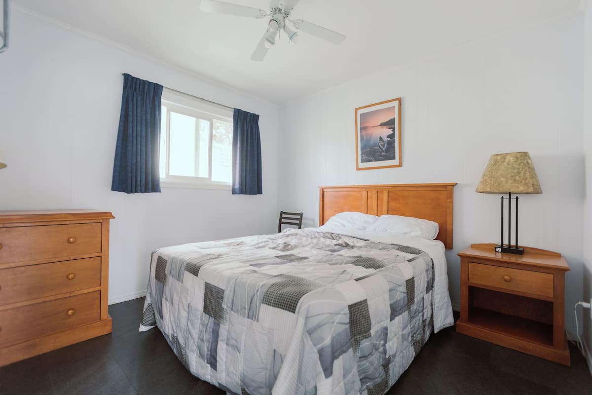 A bedroom with a bed, wooden nightstand, dresser, window with curtains, and a ceiling fan.