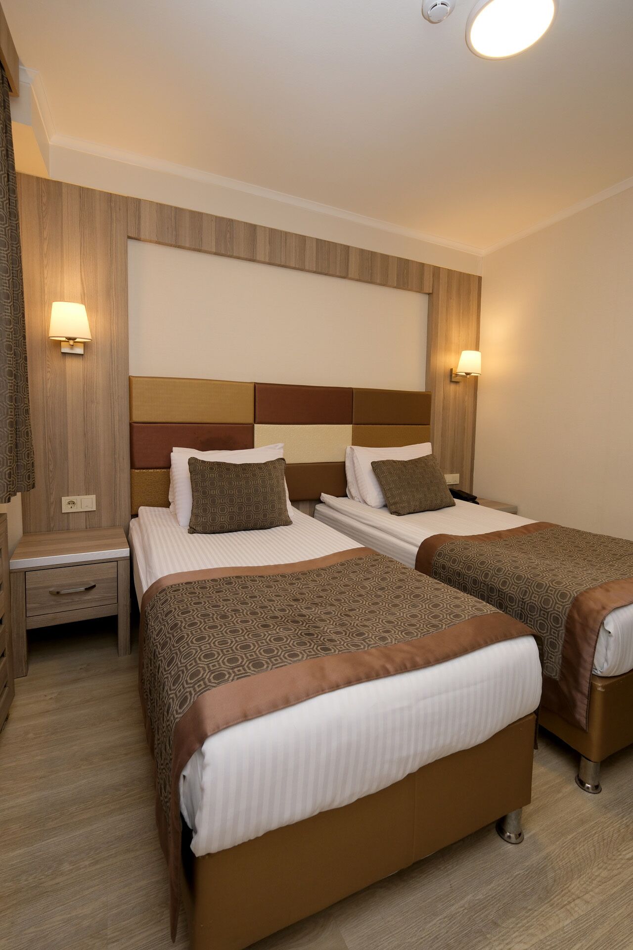 Habitación de hotel con dos camas, cabecero de madera, lámparas de noche y mesita con cajón.