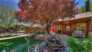 A wooden cabin with a stone waterfall feature, surrounded by a well-maintained garden with various plants and a large tree with red leaves. - Cali Cochitta Vacation Rentals (Moab)
