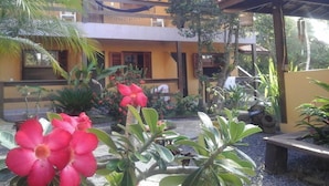 Un edifici de dues plantes amb un balcó, envoltat de plantes tropicals i una cadira espanyola.