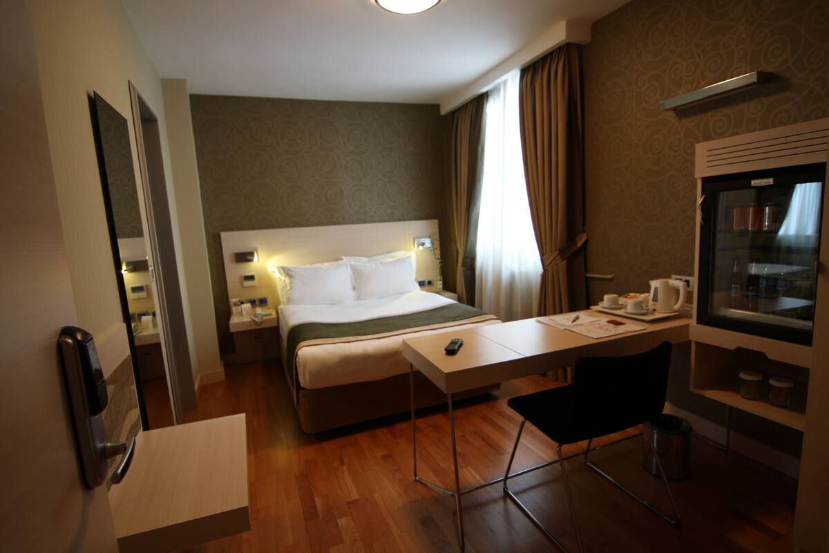 Une chambre d’hôtel comprenant un lit, un bureau, une chaise, un miroir, une fenêtre avec des rideaux et un minibar.