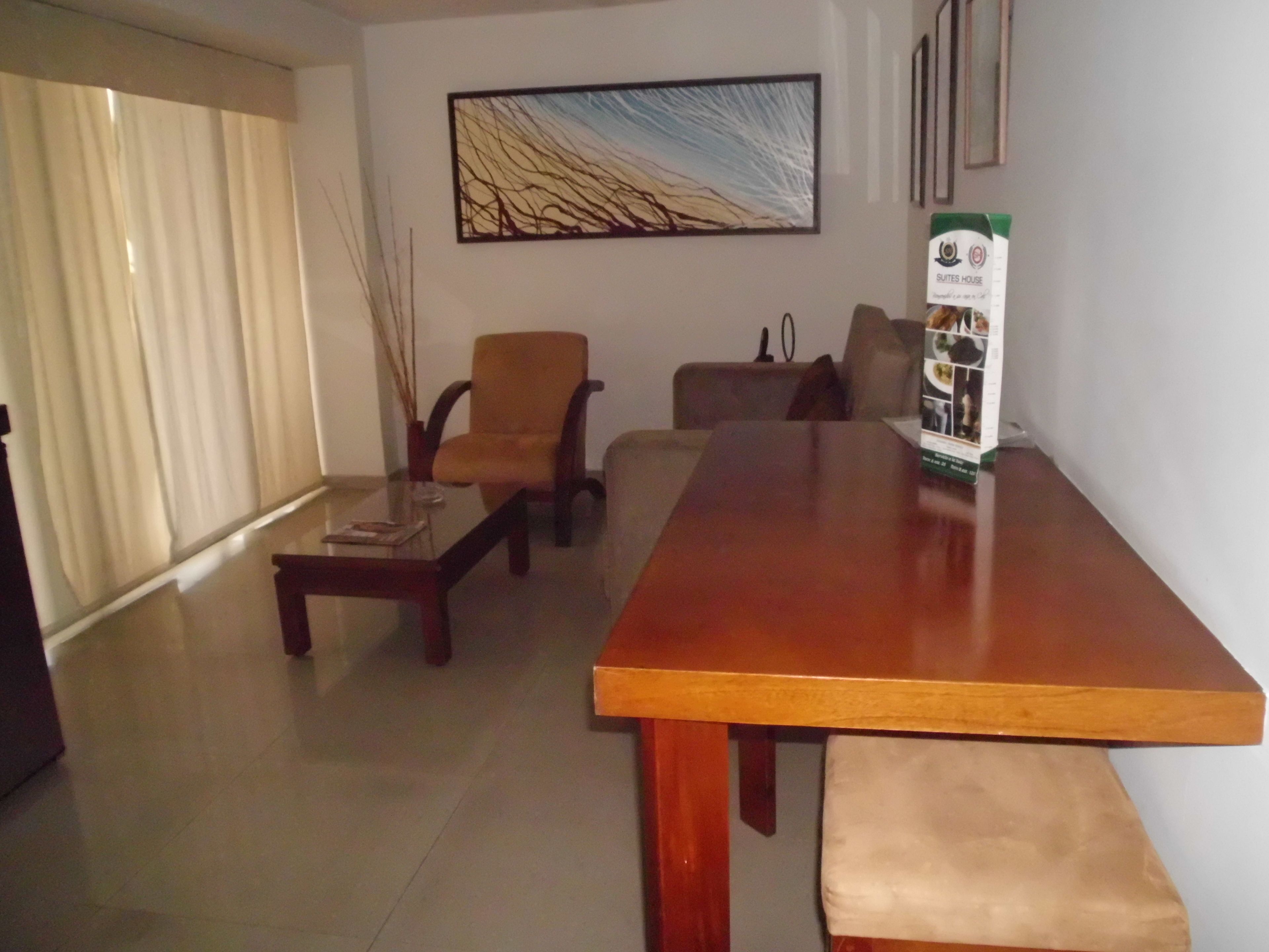Amplia sala de estar con una larga mesa de madera, dos sillones, un sofá y una mesa de centro de vidrio. Un cuadro adorna la pared, y hay un cartel sobre la mesa.
