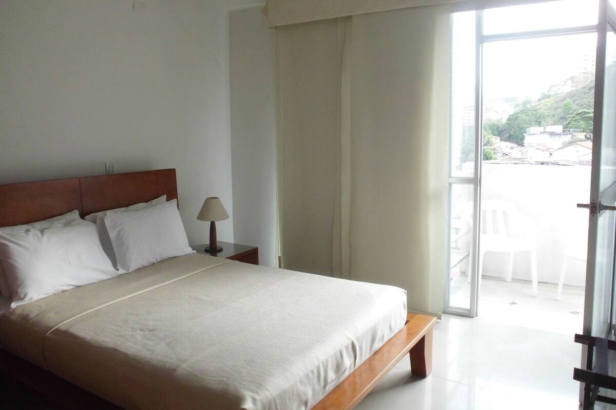 Habitación de hotel con una cama grande, una mesita de noche con lámpara y vistas al exterior a través de una puerta corrediza.