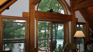Una cabaña de madera con amplios ventanales que dan a un lago, una terraza con asientos exteriores y una lámpara a la derecha.