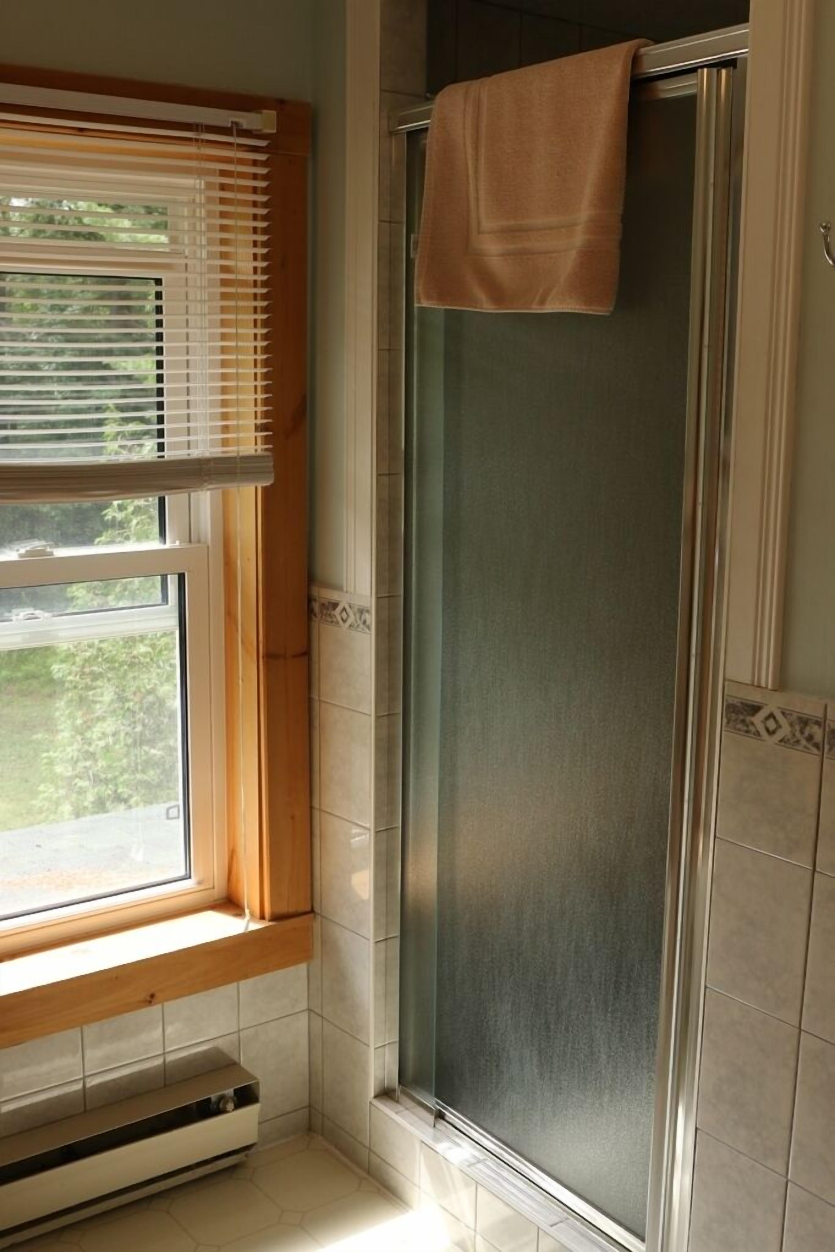 Une salle de bain avec une douche, une fenêtre avec des stores et une serviette accrochée à la porte de la douche.