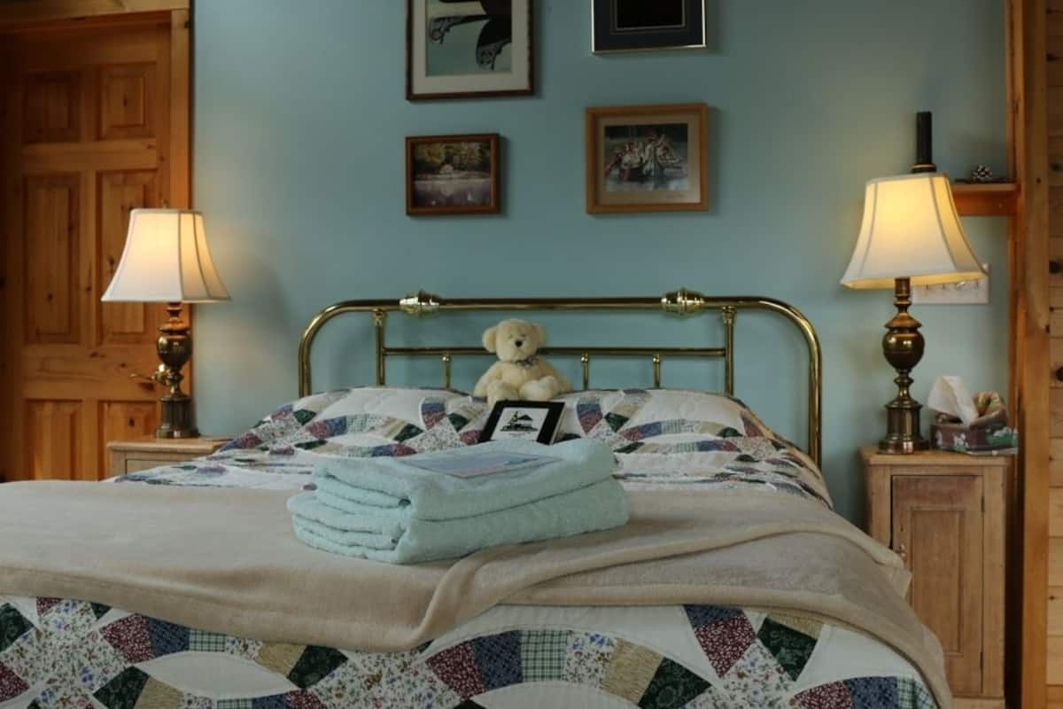 Un lit bien fait, avec une courtepointe, un ours en peluche et une serviette pliée. On y trouve deux lampes de chevet, une tête de lit en bois et un mur orné de tableaux.