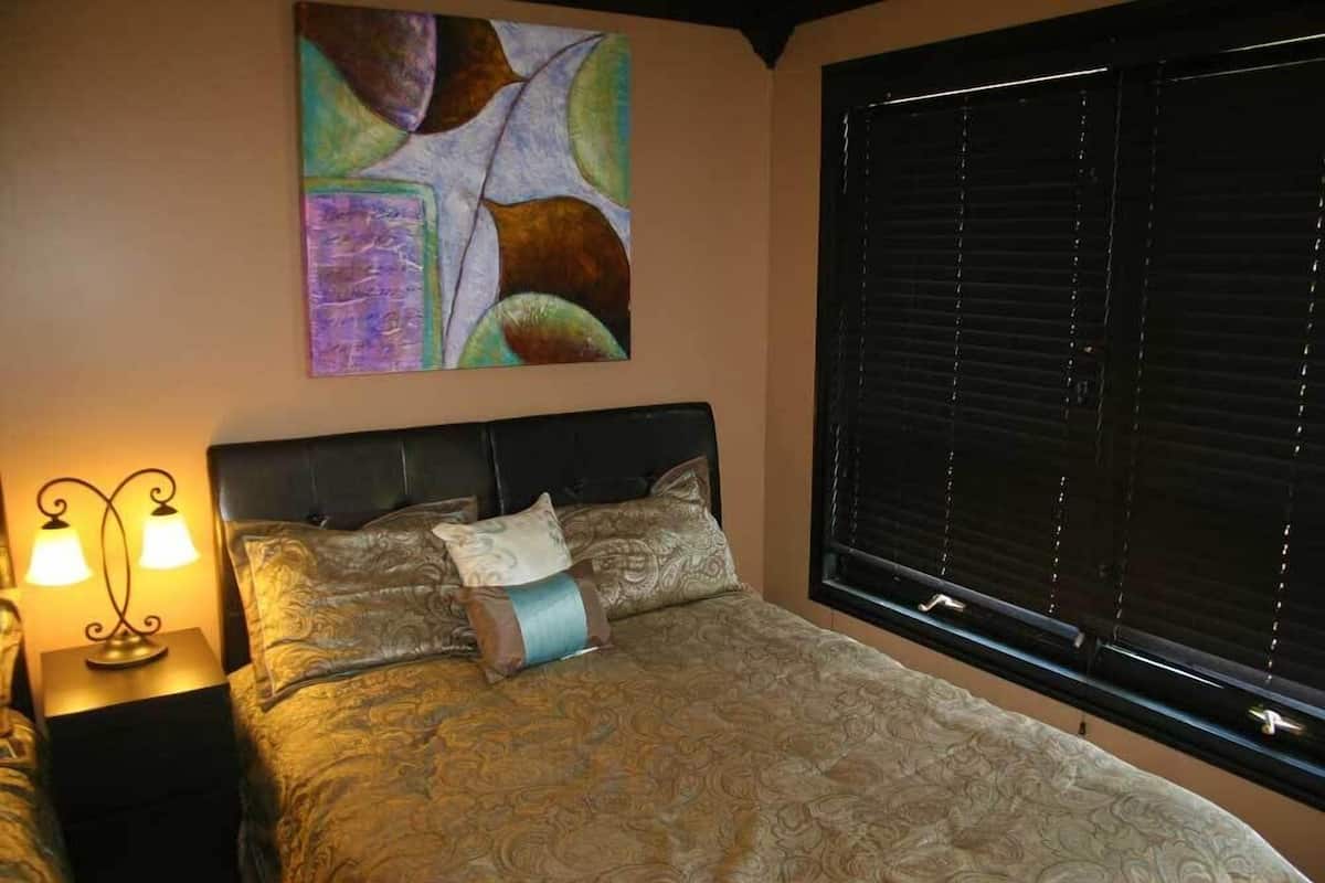 A bedroom with a large bed, a headboard, a nightstand with two lamps, and a window with blinds.