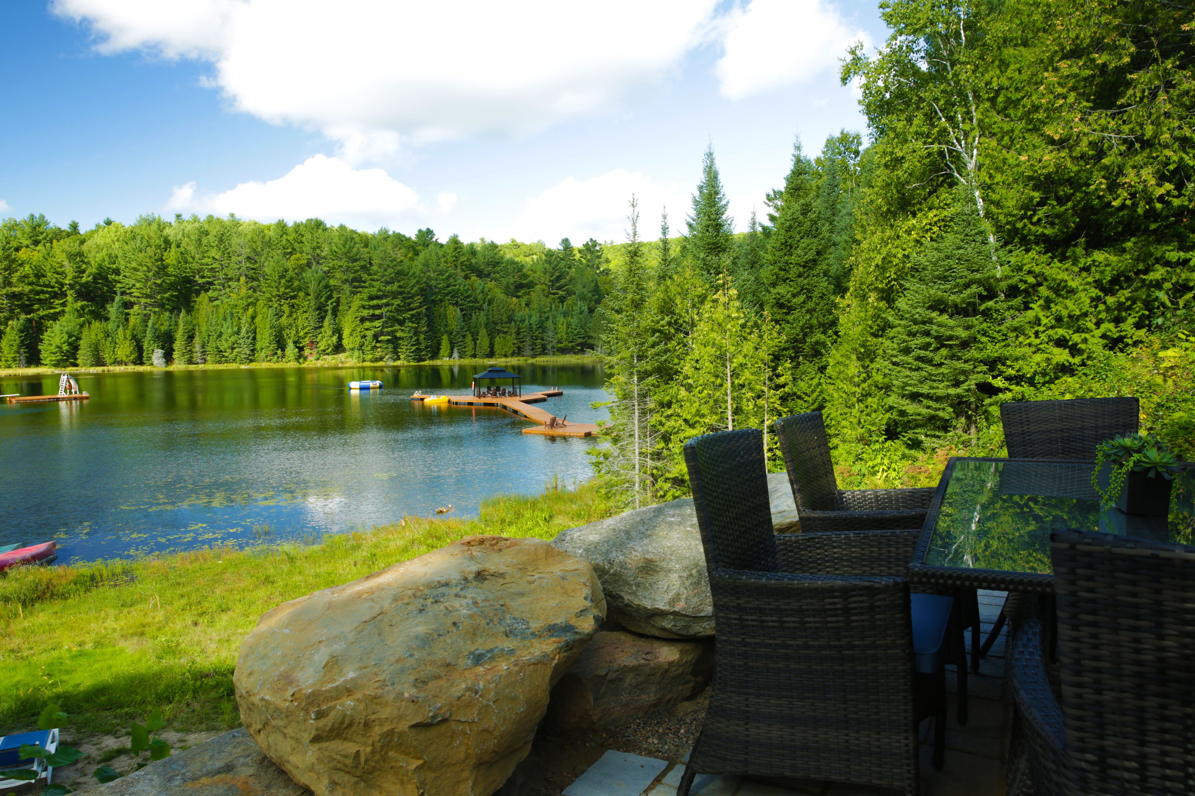 A lakeside outdoor seating area with wicker furniture and a glass table, overlooking a calm lake surrounded by dense forest.