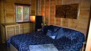 A wooden cabin room with a bed, a nightstand, a lamp, and a window with blinds. - Footprints Resort (Hastings Highlands)
