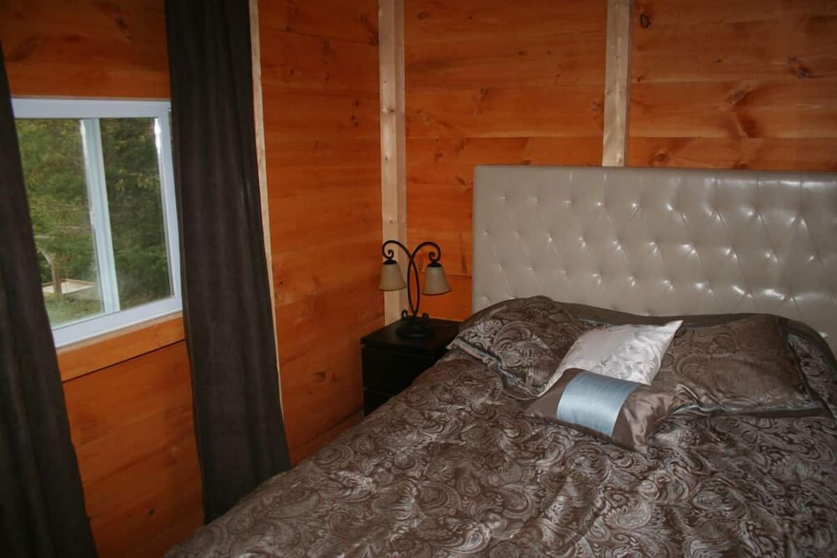 A wooden cabin room with a bed, a nightstand with two lamps, and a window with curtains.