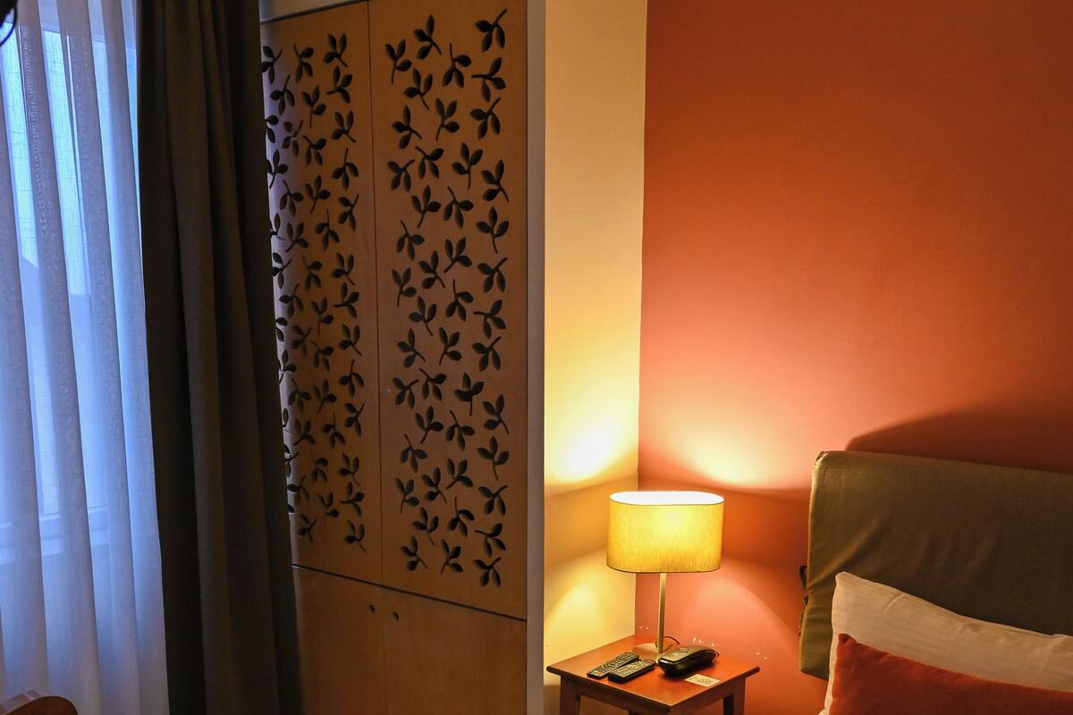 A hotel room with a bed, a nightstand with a lamp, a chair, a wooden floor, and a decorative panel.