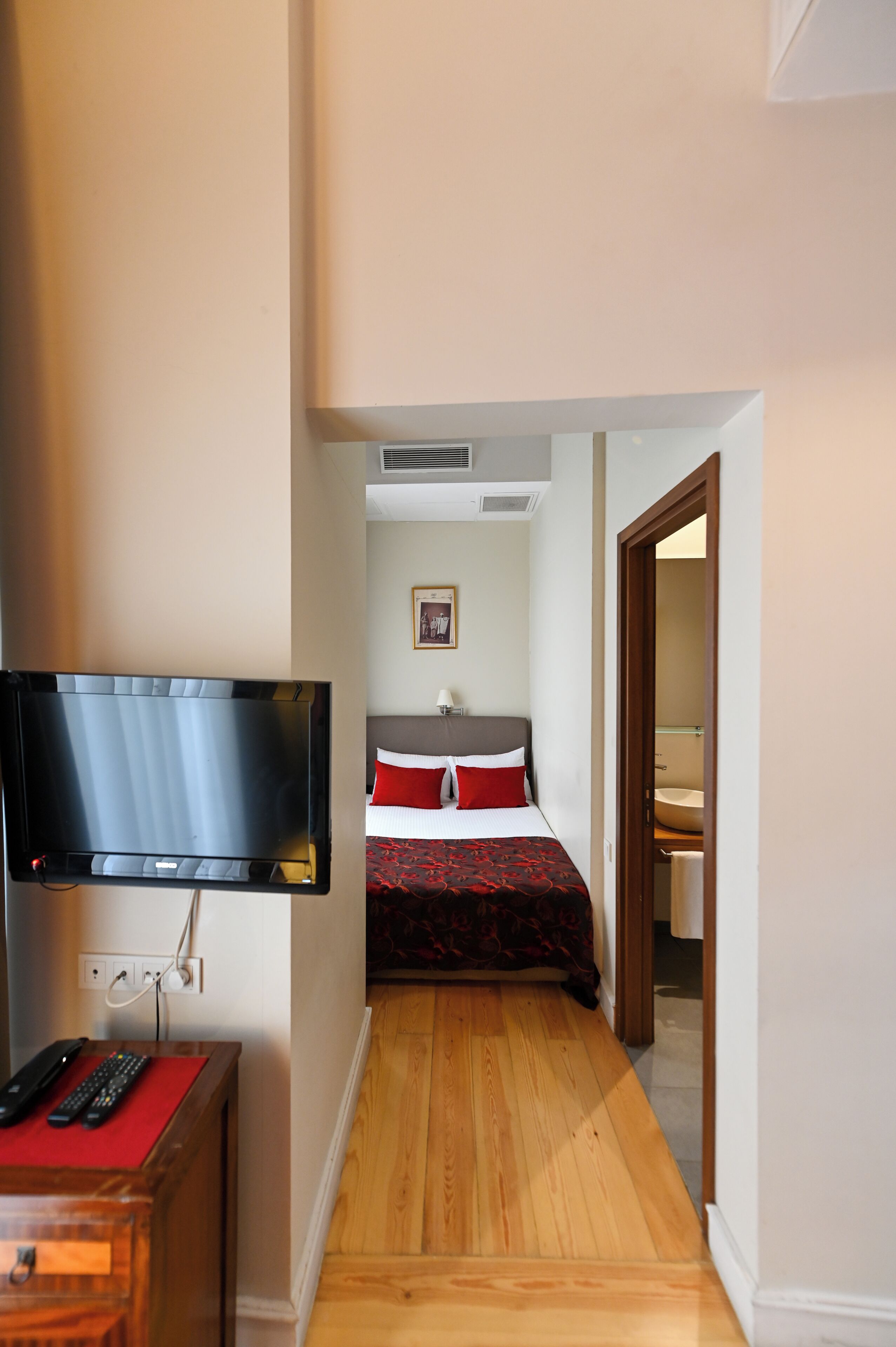 Ein Hotelzimmer mit Fernseher, einem Bett mit rot-schwarzer Bettwäsche, Holzboden und einem kleinen Holztisch mit roter Tischdecke.