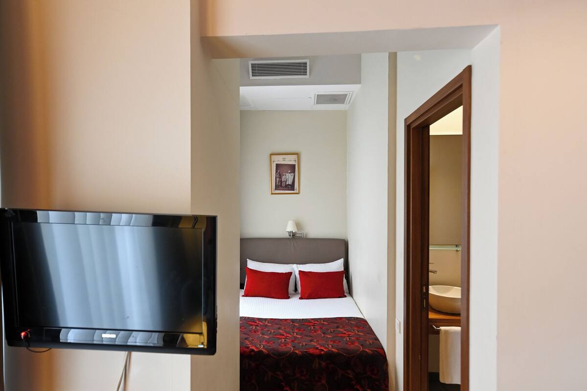 A hotel room with a television, a bed with red and black bedding, a wooden floor, and a small wooden desk with a red cloth.