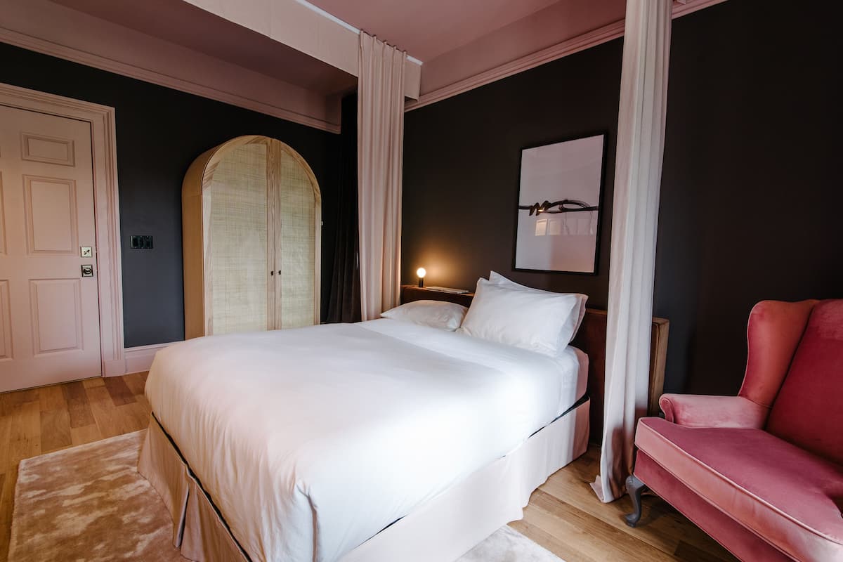 A bedroom with a large bed, a red chair, a wardrobe, and a wall-mounted artwork.