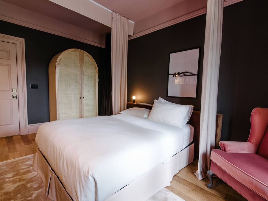 Une chambre avec un grand lit, un fauteuil rouge, une armoire et une œuvre d’art accrochée au mur.
