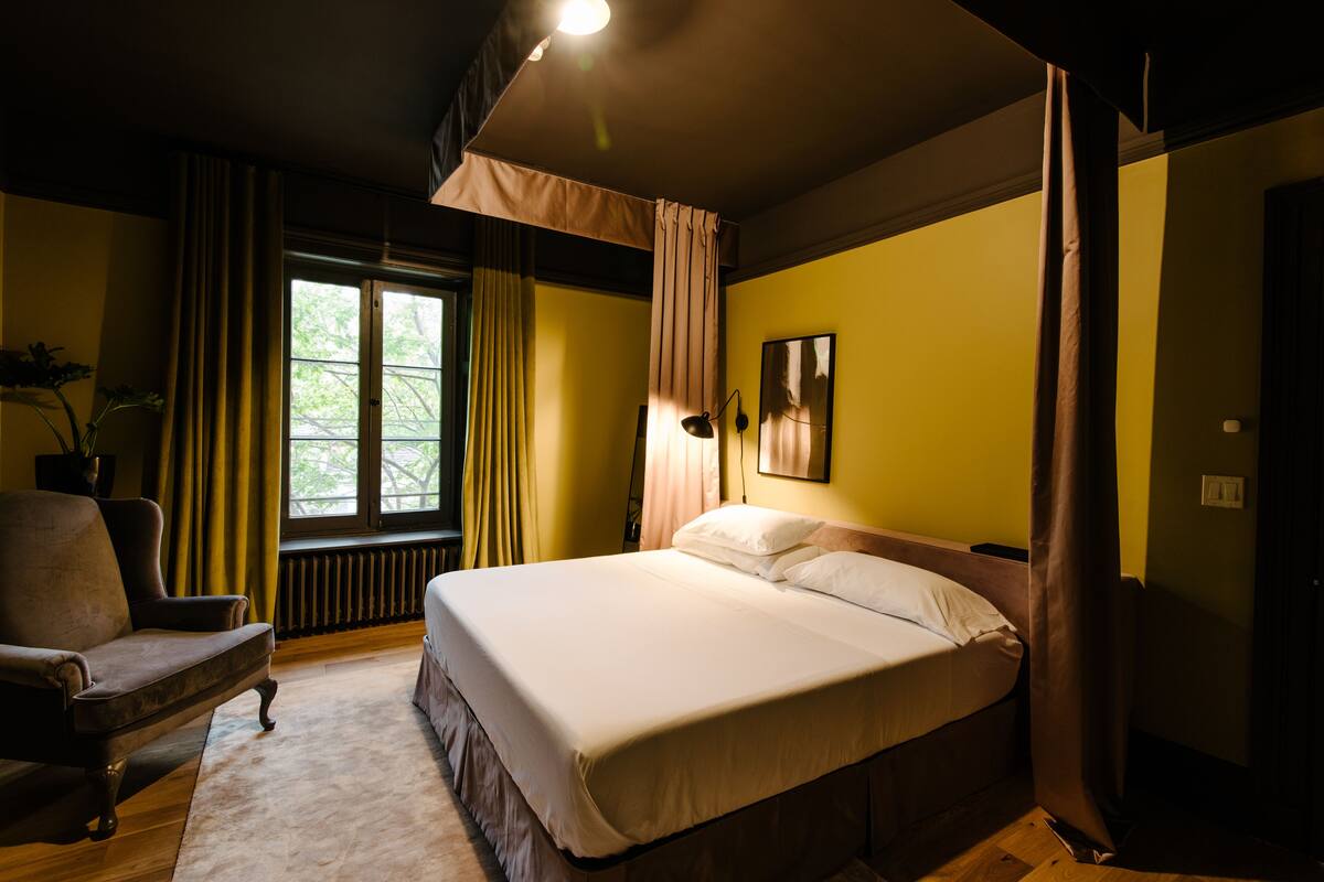 A bedroom with a bed, a chair, a window with curtains, and a radiator.