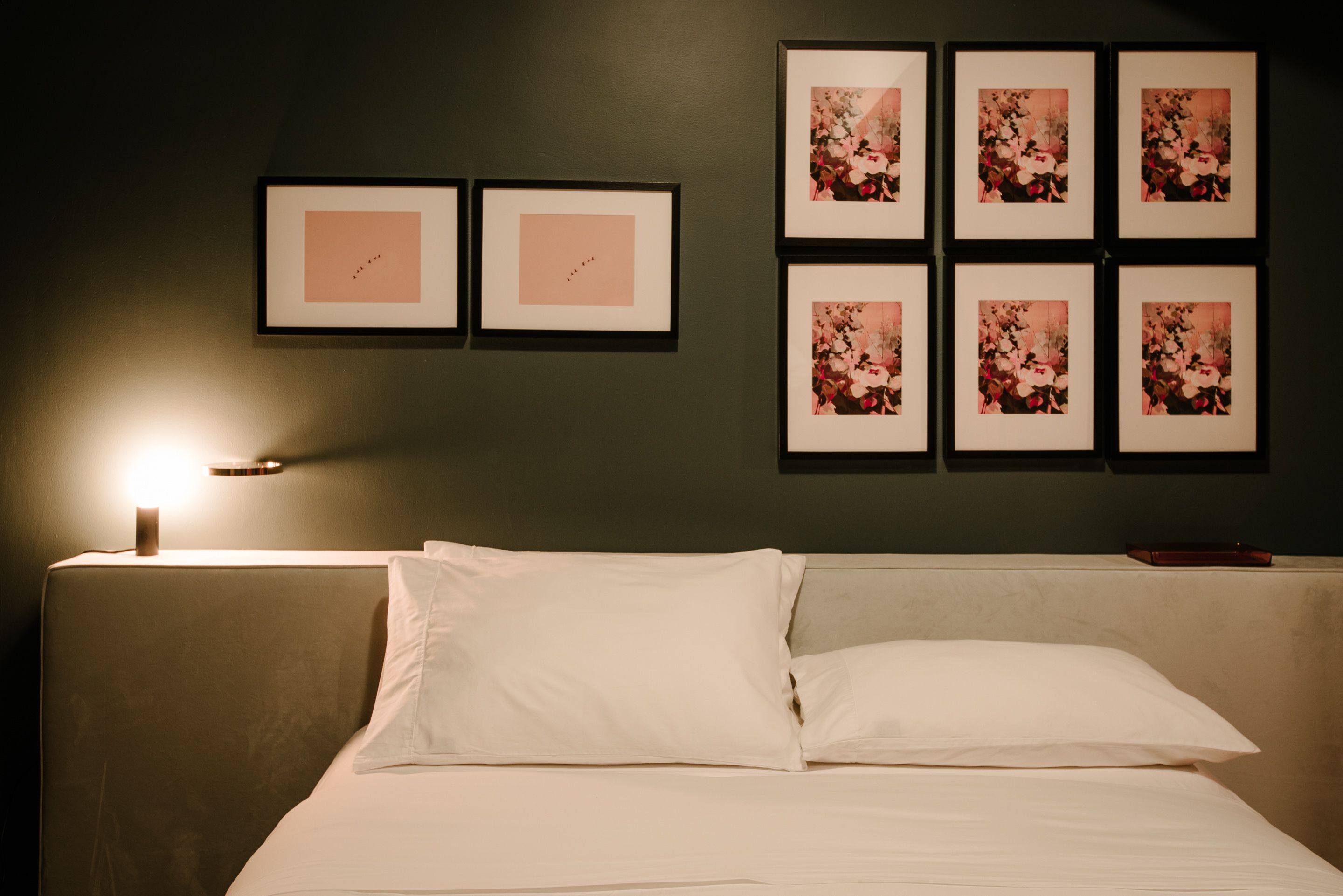 A neatly made bed with white linens, a bedside table with a lamp, and six framed floral pictures on the wall.