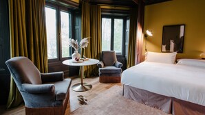 Hotelkamer met een groot bed, twee fauteuils, een kleine ronde tafel met een vaas, en uitzicht op bomen door de ramen.