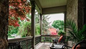 Een overdekt terras met uitzicht op een tuin en een bakstenen gebouw. Twee stoelen en een tafel staan er, met een vloerkleed.