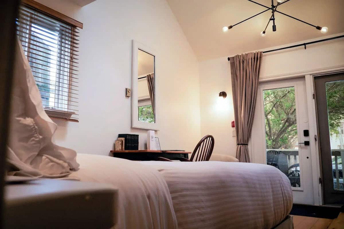A bedroom with a bed, a desk with books, a mirror, and a view of the outdoors.