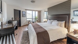 A modern hotel room with a large bed, a desk with a chair, a small table, and a view of the city through large windows. - Jose Antonio Executive (Lima)