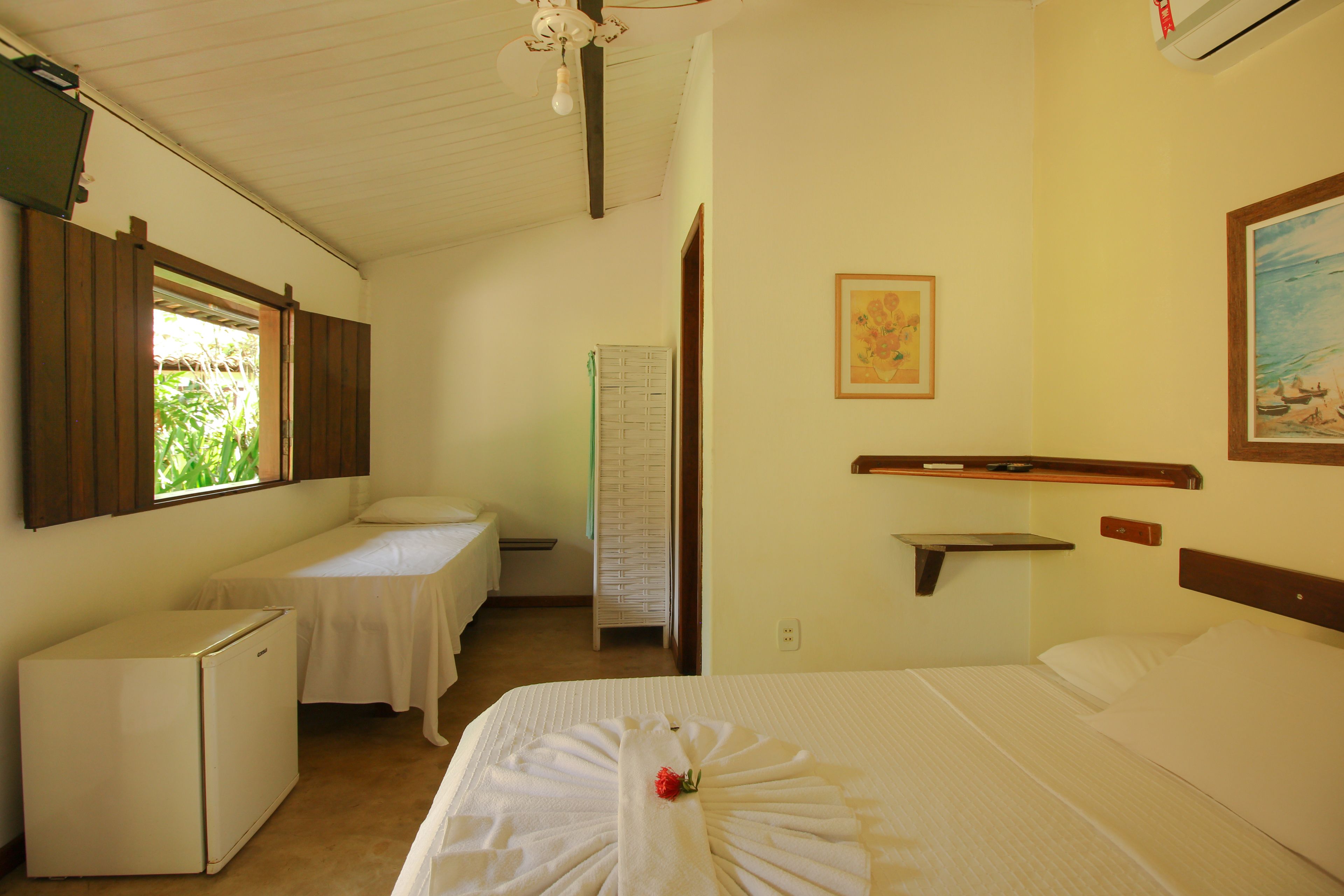 Quarto de hotel com uma cama, um pequeno frigorífico, uma escrivaninha e uma janela com persianas.