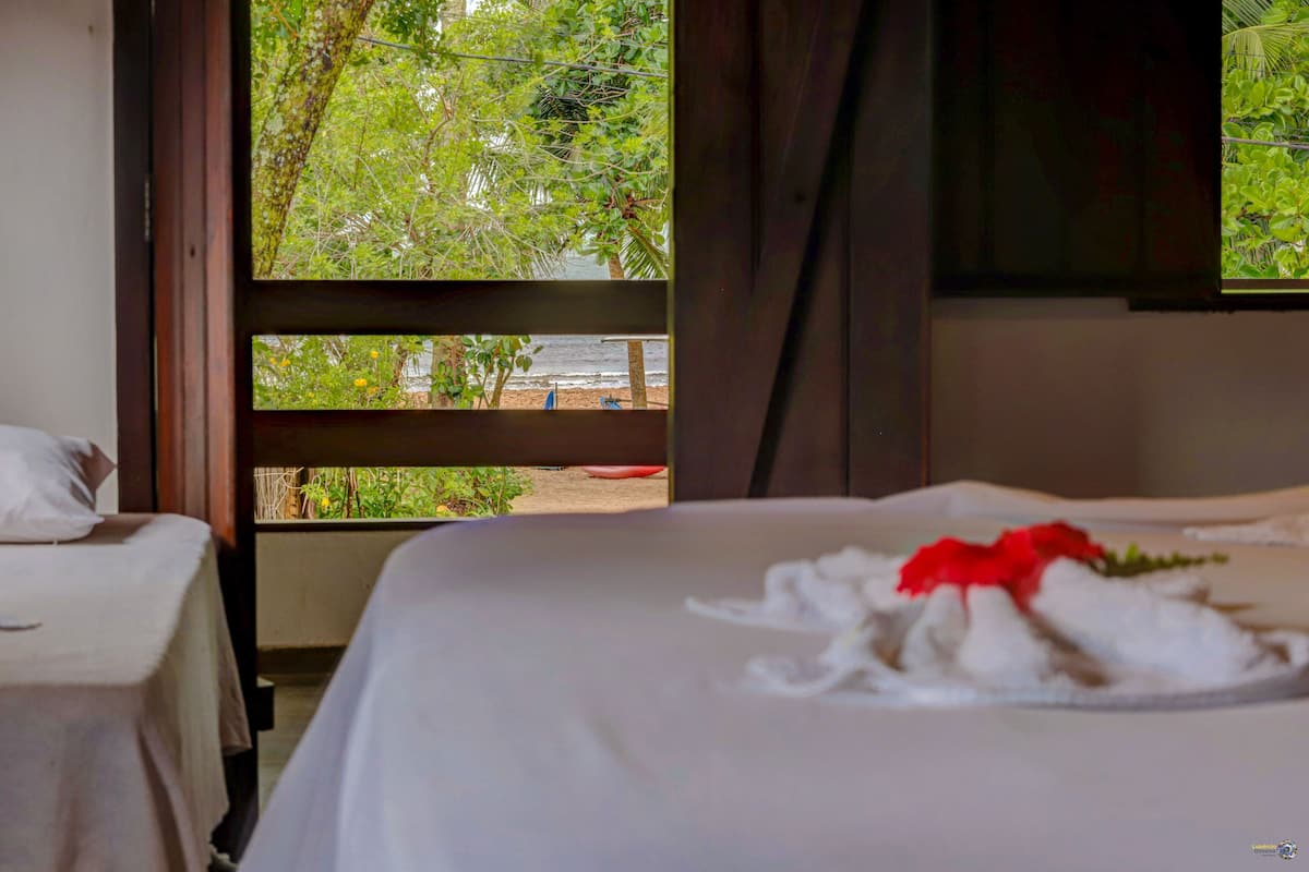 A hotel room with a bed, a window with a view of greenery and a beach, and a wooden railing outside.