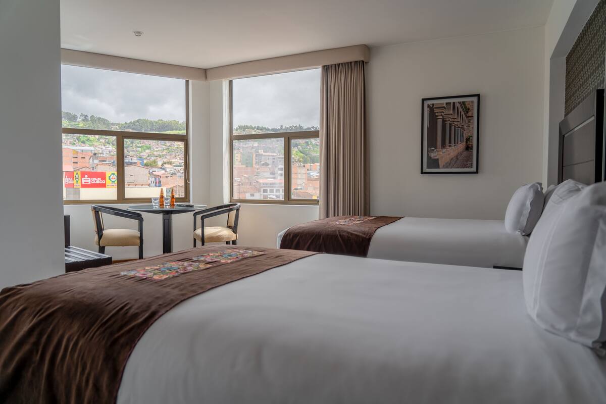 Quarto de hotel com duas camas, uma escrivaninha e uma cadeira. Há uma janela grande com vista para edifícios e paisagem urbana.